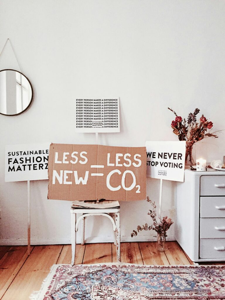 Cardboard and paper protest-style signs with eco-conscious slogans like “LESS NEW = LESS CO₂” and “SUSTAINABLE FASHION MATTERZ” in a home interior setting