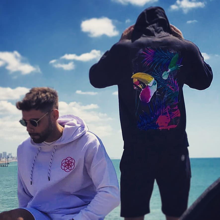 scar-tissue-beach-streetwear-photo-graphic-hoodies Two men wearing streetwear hoodies by the beach; one in a white hoodie with a geometric logo, the other in a black hoodie featuring a vibrant tropical bird and foliage print on the back.