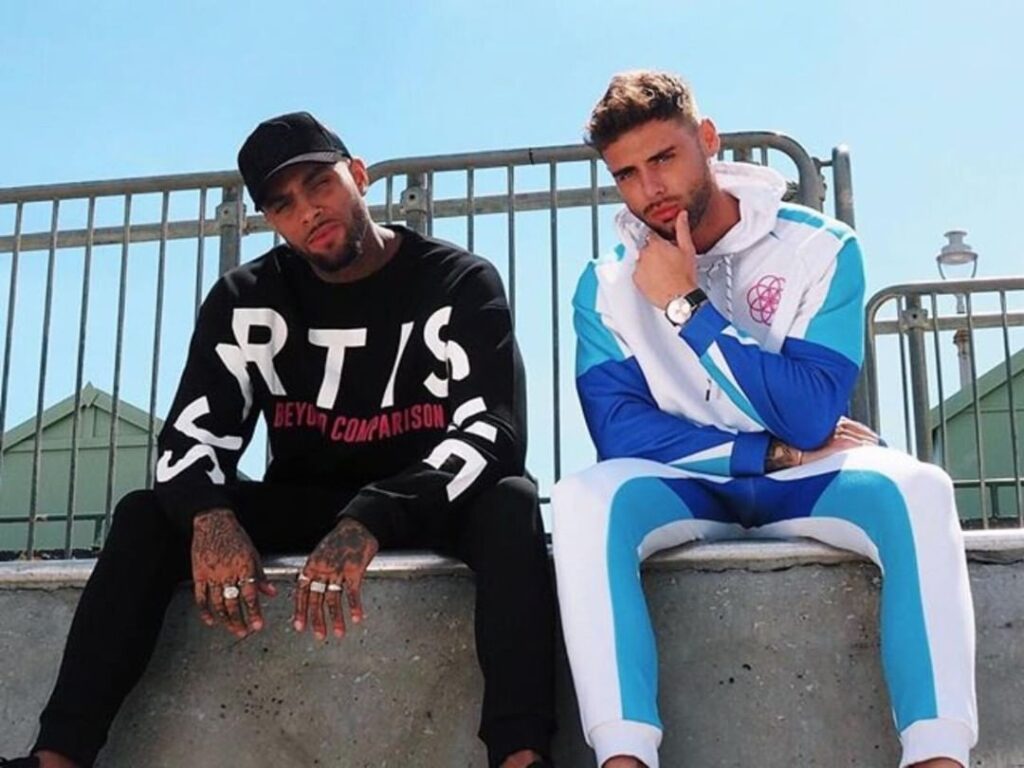 scar-tissue-black-sweater-beyond-comparison Two male models sit on a concrete ledge wearing Scar Tissue streetwear; one in a black “RTIS Beyond Comparison” sweatshirt, the other in a blue and white tracksuit with a geometric chest logo.