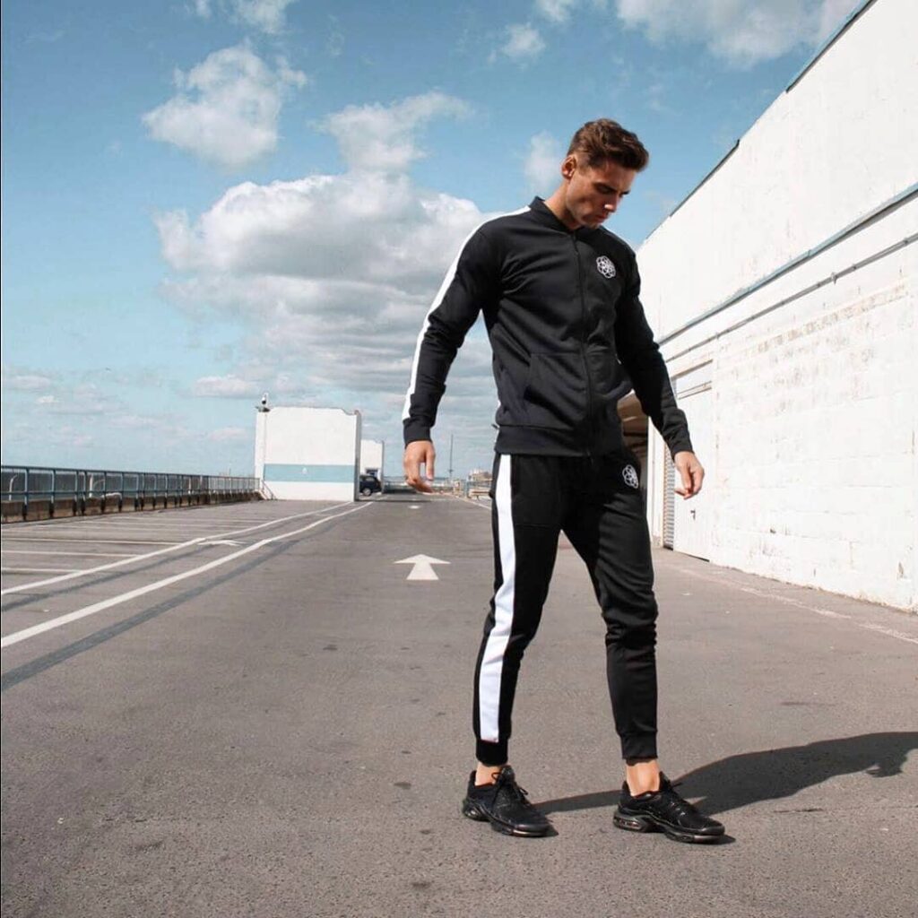 scar-tissue-black-tracksuit-seafront-streetwear Male model wears a black Scar Tissue tracksuit with white side stripes, standing on an empty seafront road under a partly cloudy sky.