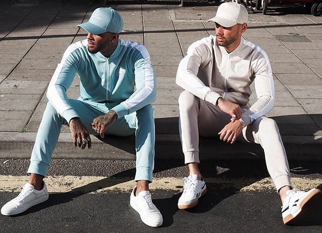 scar-tissue-blue-and-beige-tracksuits-duo Two male models wear pastel-toned Scar Tissue tracksuits—one in powder blue, one in beige—while sitting on a kerb in bright sunlight.