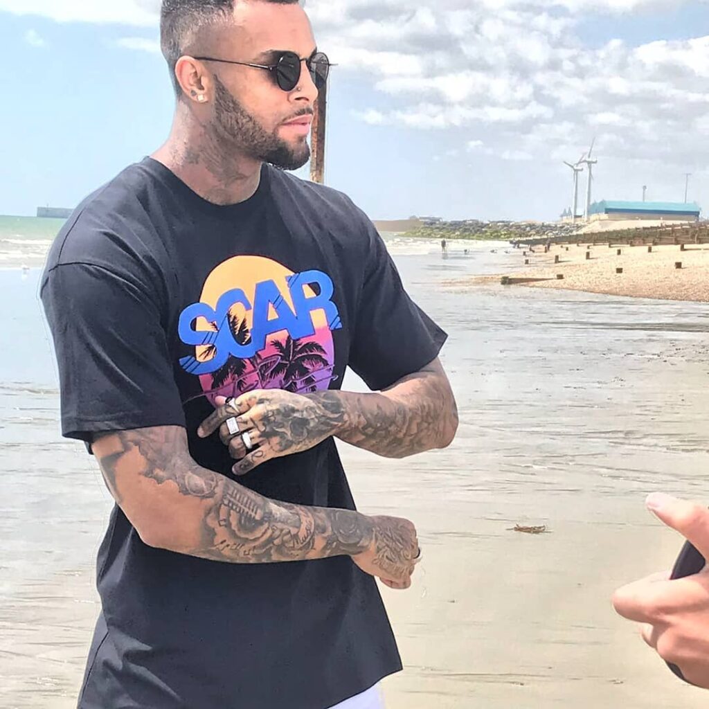scar-tissue-graphic-logo-tee-sunset-print Male model wearing a black Scar Tissue T-shirt with a bold sunset graphic and palm tree print, standing on a beach with wind turbines in the distance.