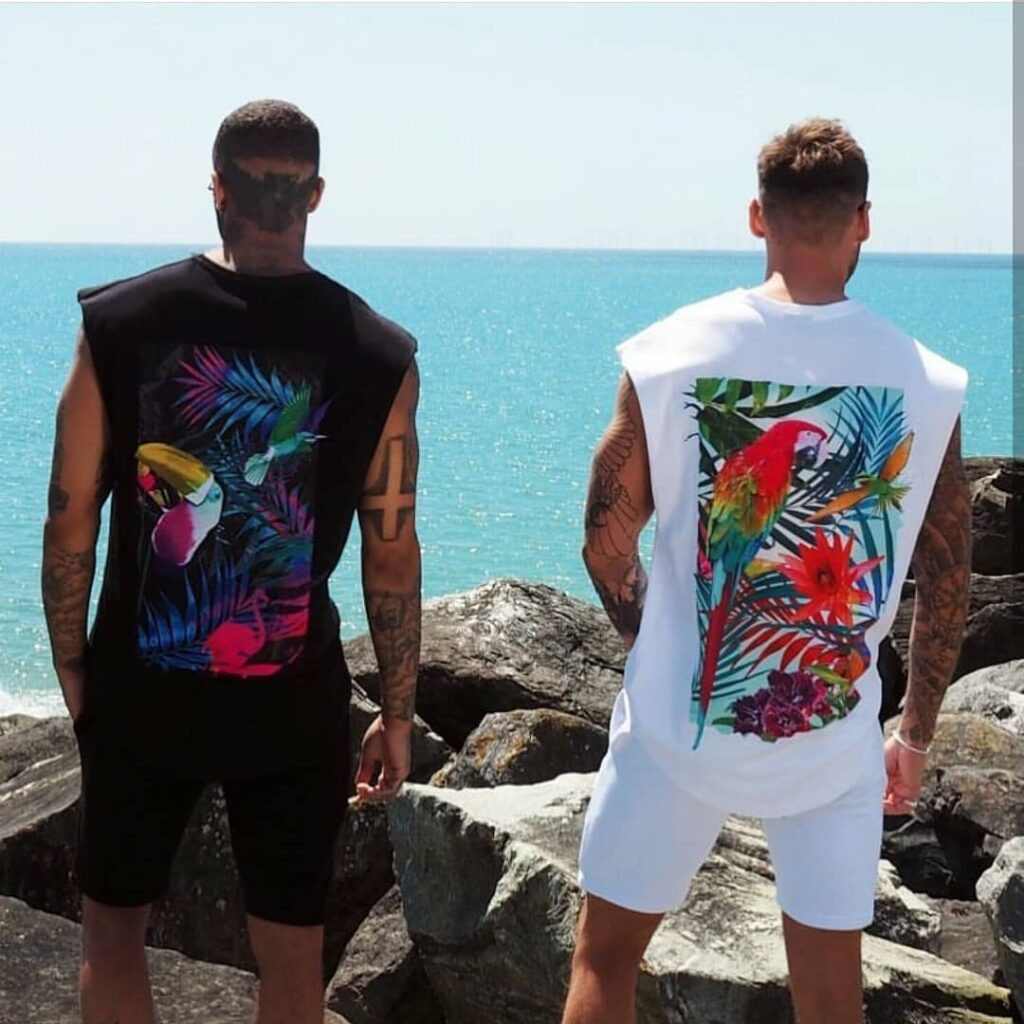 scar-tissue-parrot-graphic-sleeveless-beachwear Two male models wear sleeveless Scar Tissue tops with vibrant parrot and jungle graphics, standing on rocks by the sea.