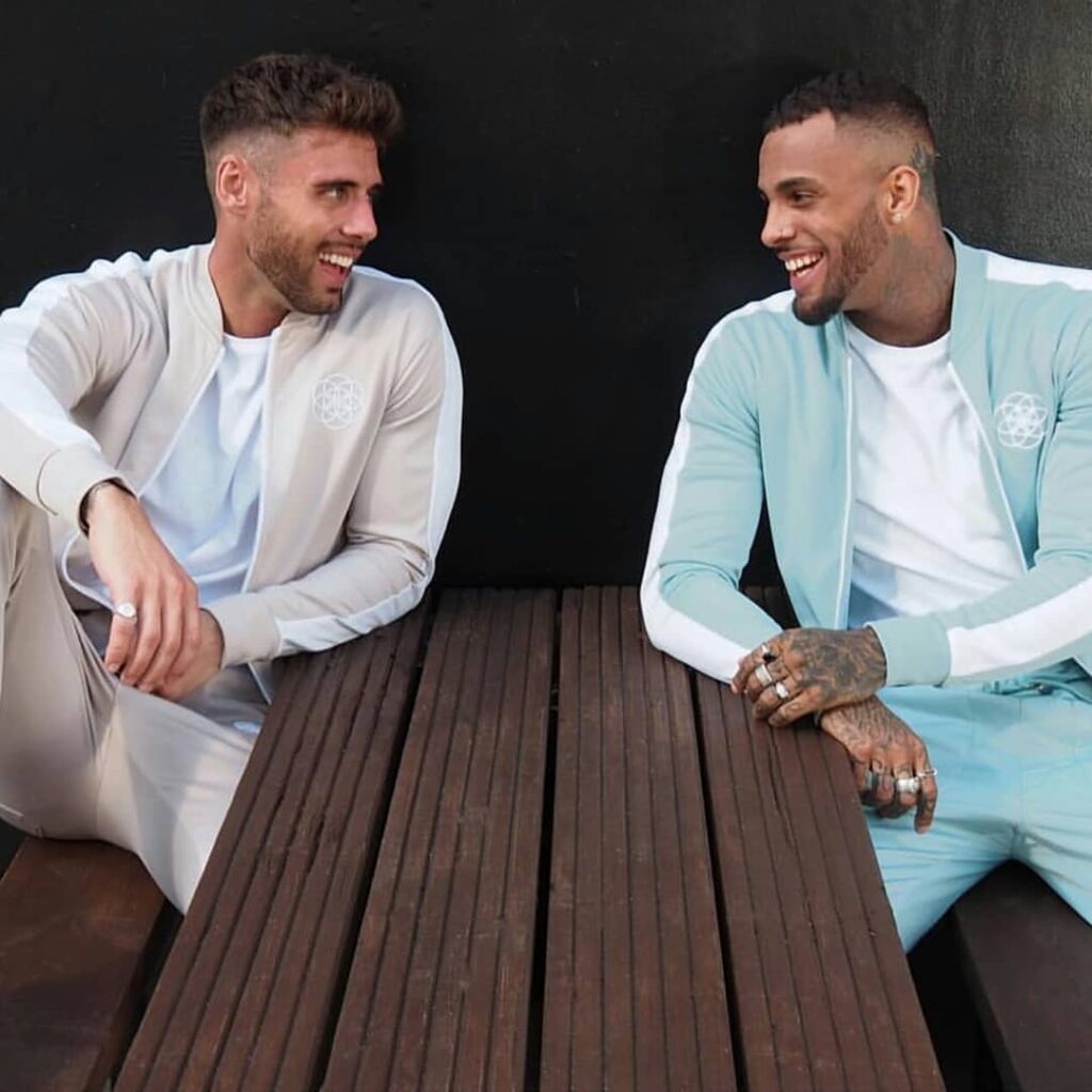 scar-tissue-pastel-tracksuits-lifestyle-fashion Two male models wearing pastel Scar Tissue tracksuits—one in beige, one in mint—sit at a wooden bench, laughing together.