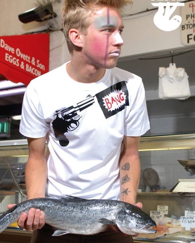 sloth clothing brighton mens streetwear designer Male model in a white Sloth T-shirt with gun and “BANG” graphic holding a fresh fish in the London Road Market Brighton fish shop