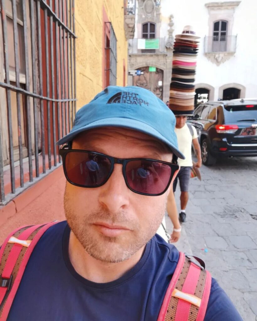 Jonathan Mitchell wearing sunglasses and a North Face cap, with a street vendor behind him balancing a tall stack of hats on his head – representing the metaphor of wearing many hats
