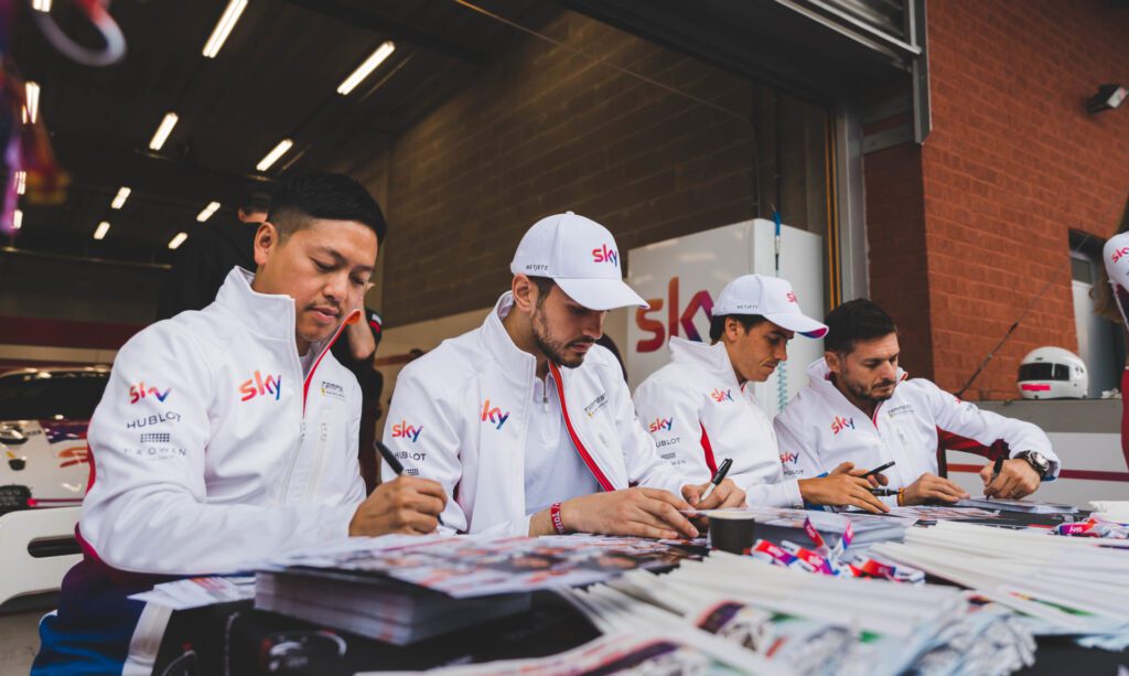 4 members of the Sky Tempesta GT3 motorsport team wearing white branded teamwear jackets and caps designed by Jonathan Mitchell, signing autographs at a race event.