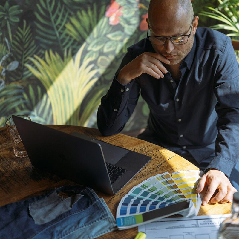 Jonathan Mitchell, freelance fashion designer, reviewing colour swatches and denim samples while working at a laptop in a vibrant, plant-filled workspace.